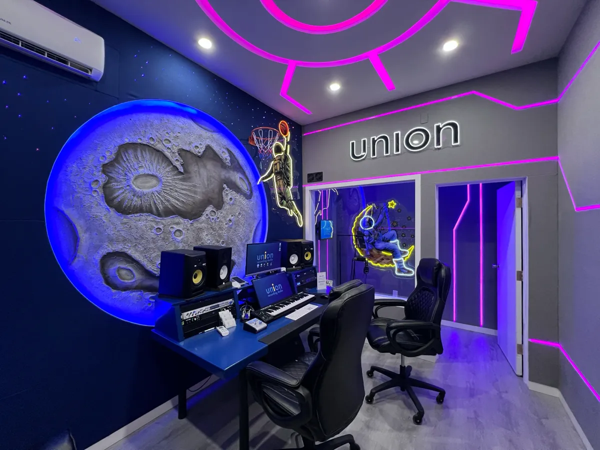 COSMO room at UNION: desk with studio monitors, a screen and a keyboard controller, a sculpted moon relief lit blue, and an astronaut neon sign.