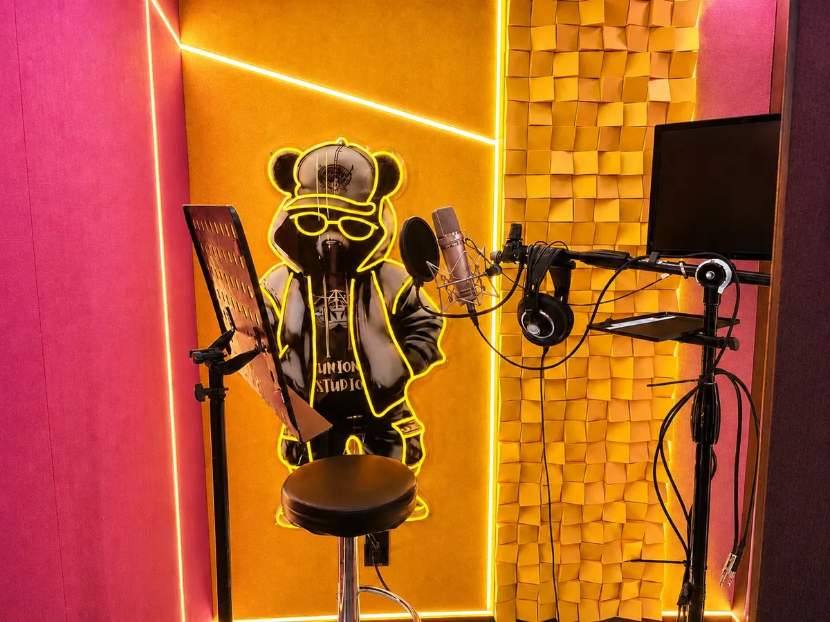 LEGACY vocal booth at UNION: condenser microphone and pop filter on a boom stand, headphones, a music stand and a screen beside orange acoustic diffusers.